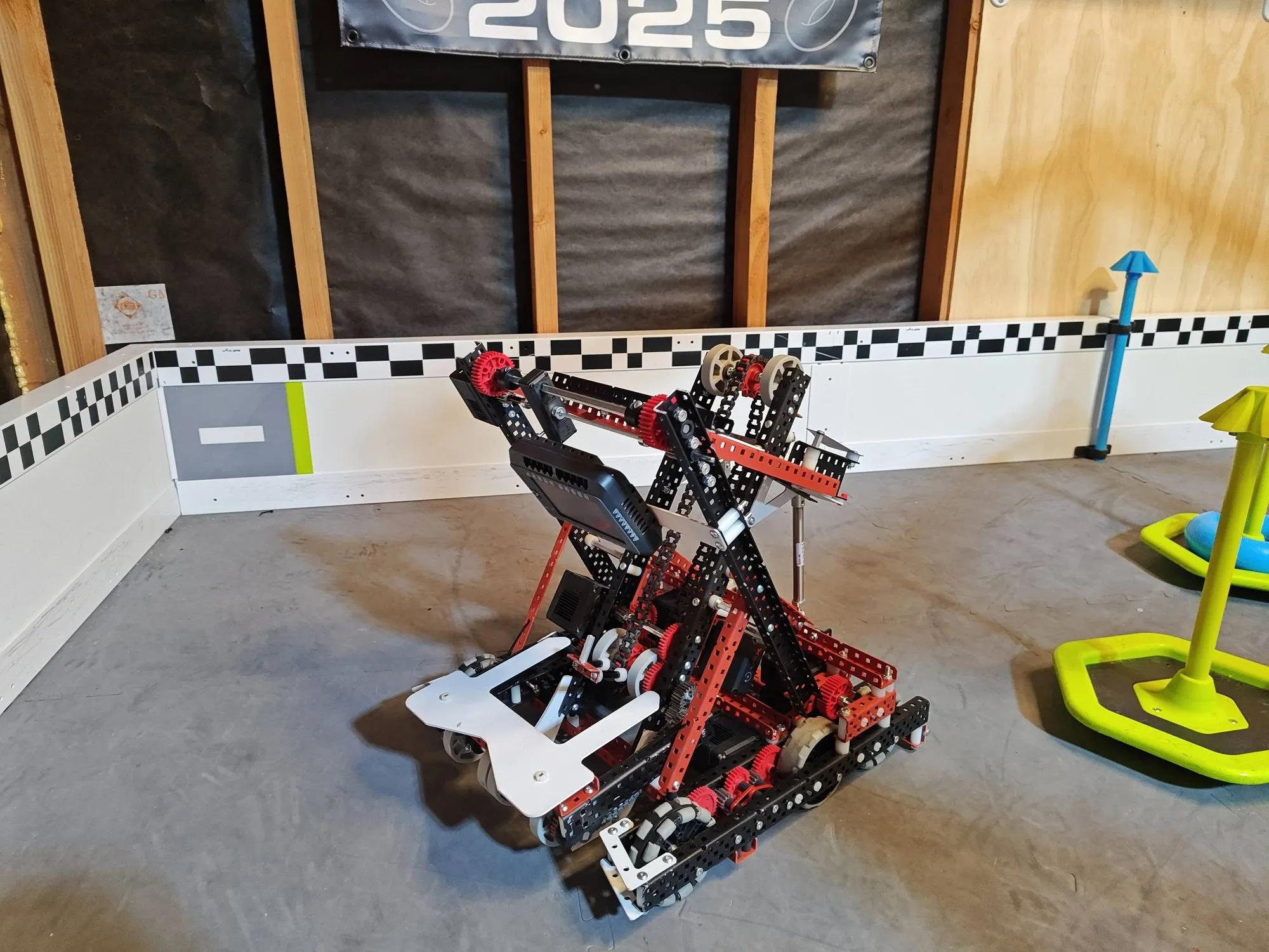 The High Stakes robot, a red and black frame with a tall lift, on a practice field with checkered walls.
